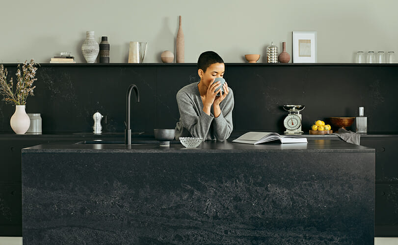 אישה שותה קפה ומניחה את המרפקים במטבח על משטח של אבן קיסר